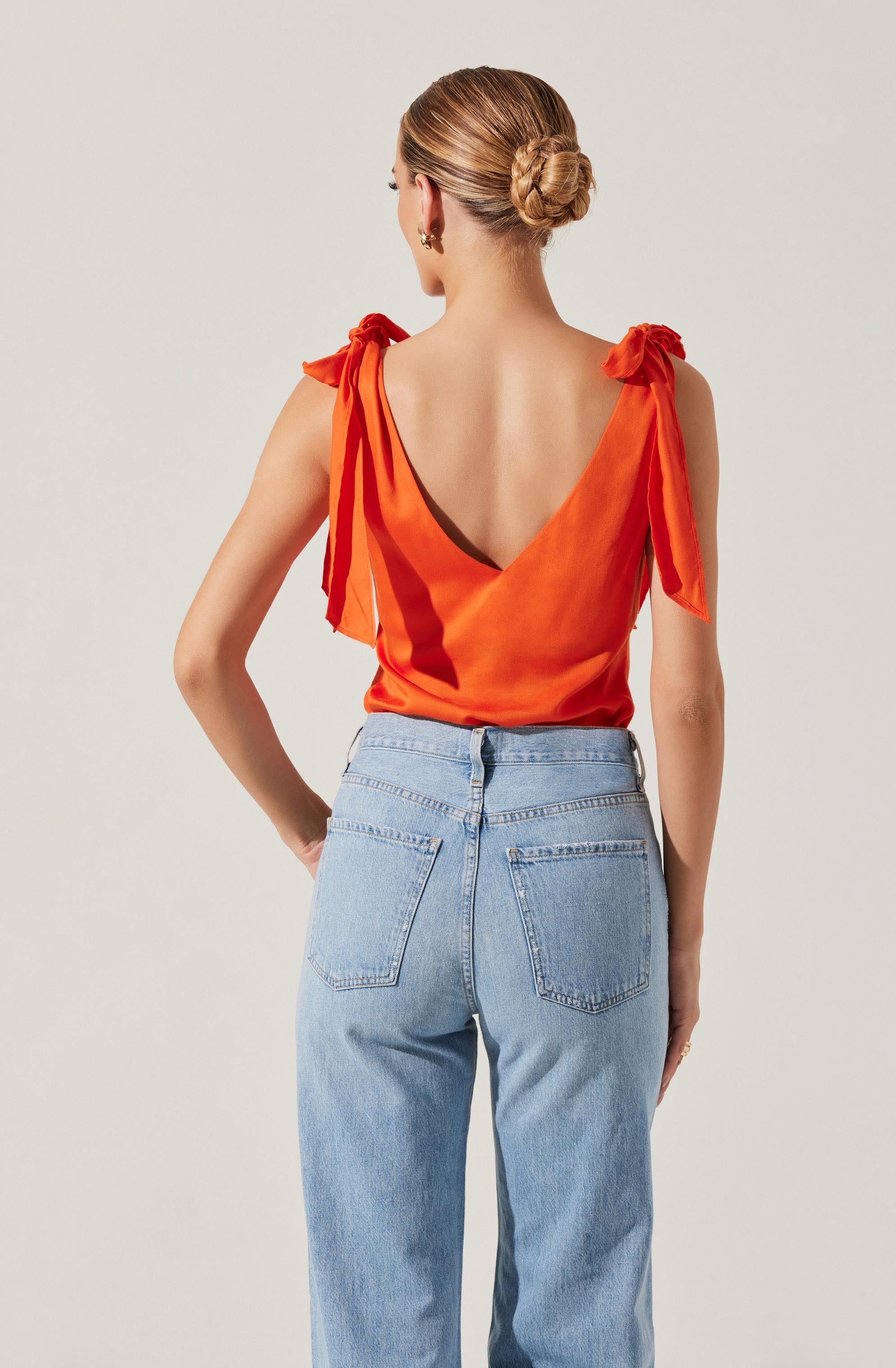 A woman is wearing a sleeveless, orange satin top with a deep V-back and tied shoulder straps. She is also wearing high-waisted, light blue denim jeans.