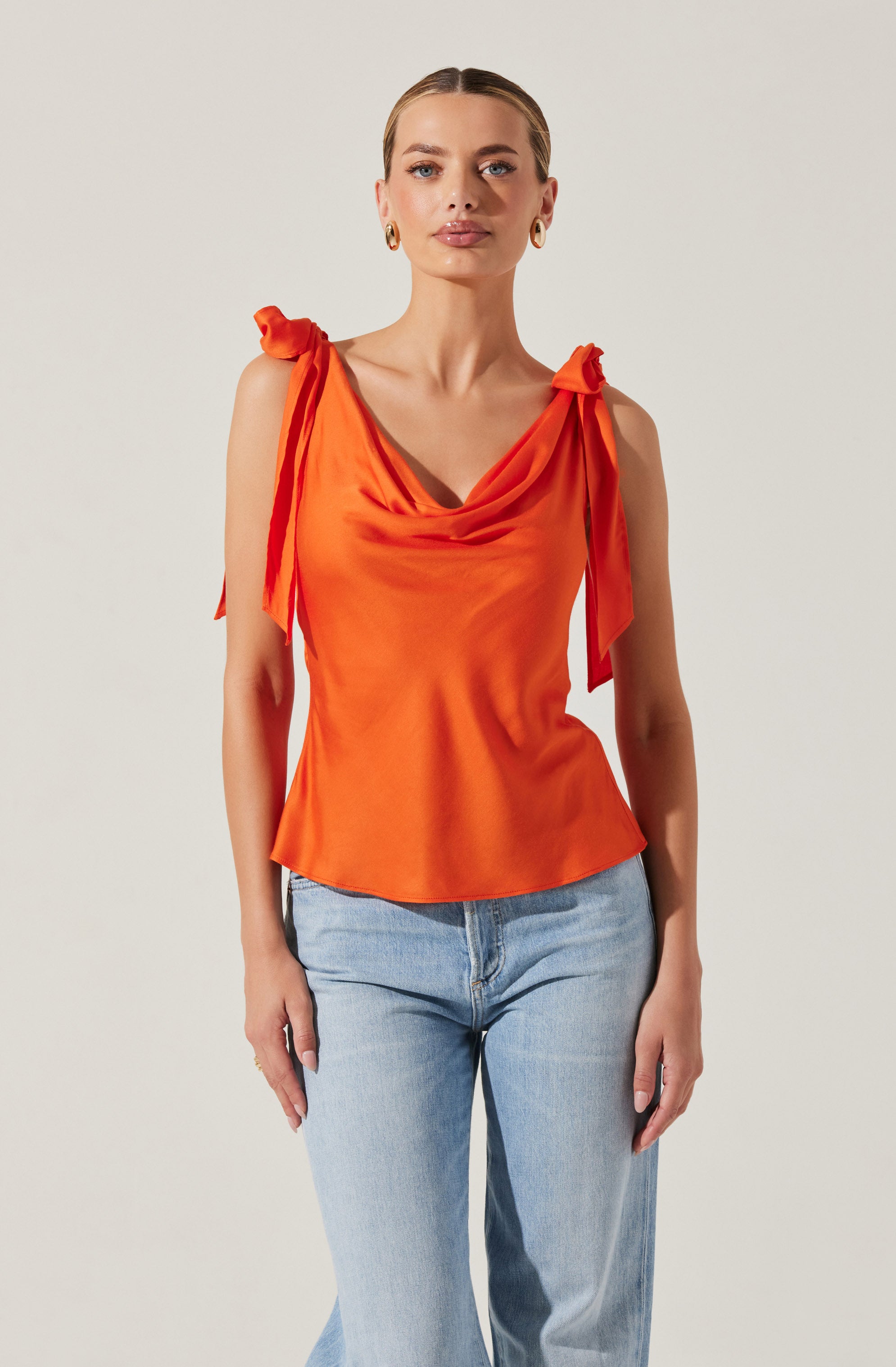 A woman is wearing a sleeveless, bright orange satin top with draped details at the neckline and shoulder ties that form bows. She pairs it with light blue high-waisted jeans.