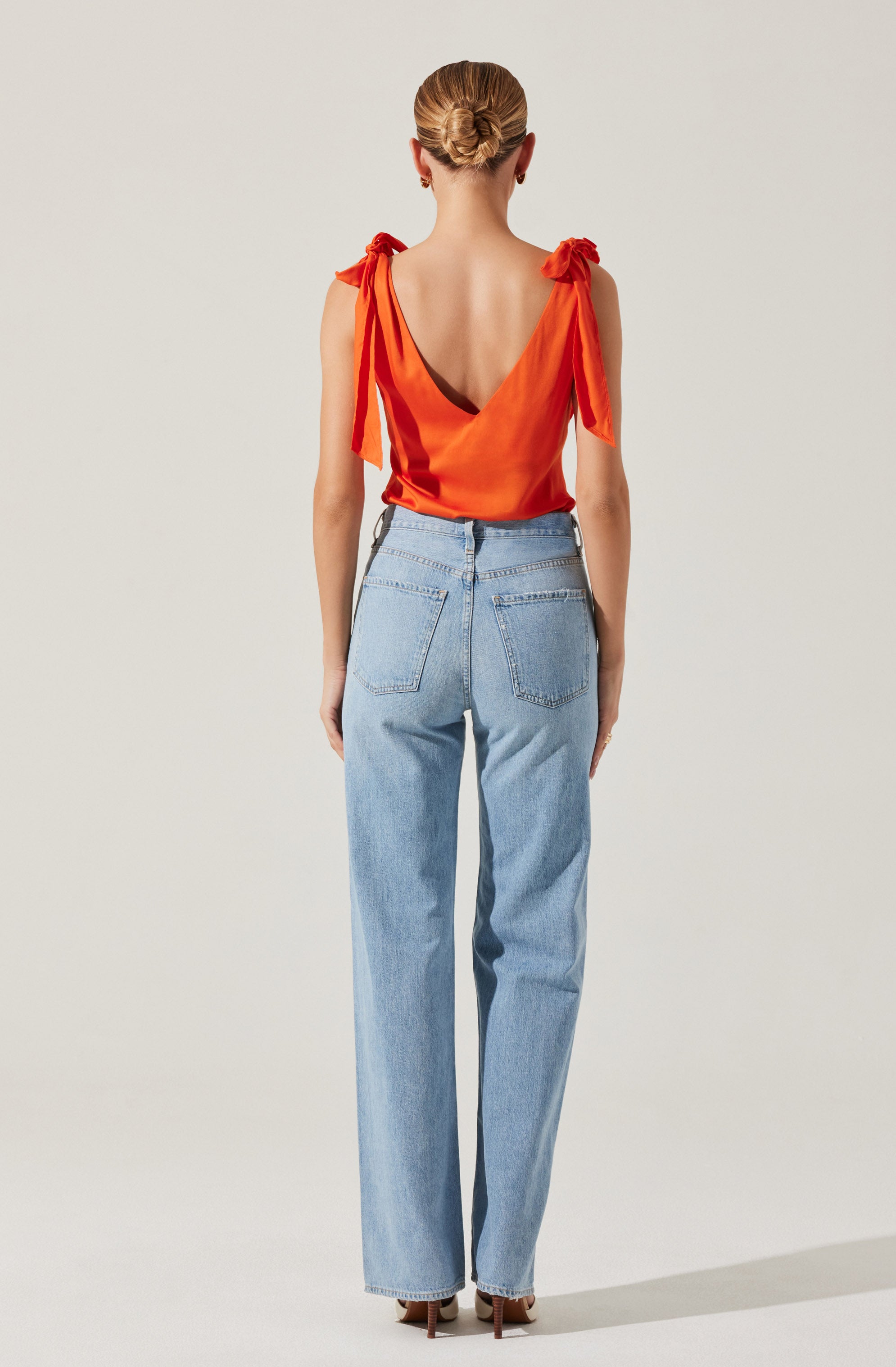A woman is wearing a sleeveless, orange satin top with shoulder tie details and a deep V-back. She is also dressed in high-waisted, light blue denim jeans with back pockets. Her hair is styled in a neat bun.
