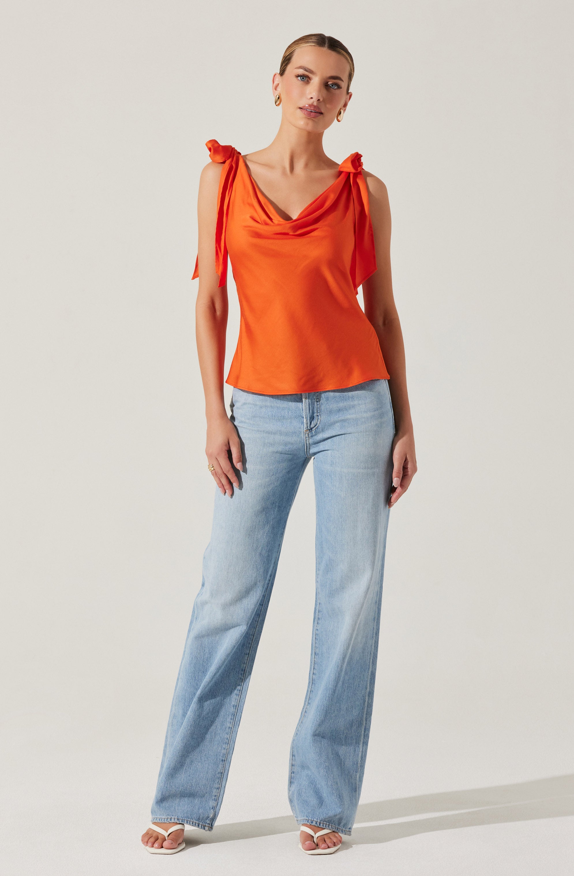 A woman is wearing a sleeveless orange satin top with shoulder ties and a draped neckline, paired with light blue high-waisted, wide-leg jeans. She is also wearing open-toe sandals.