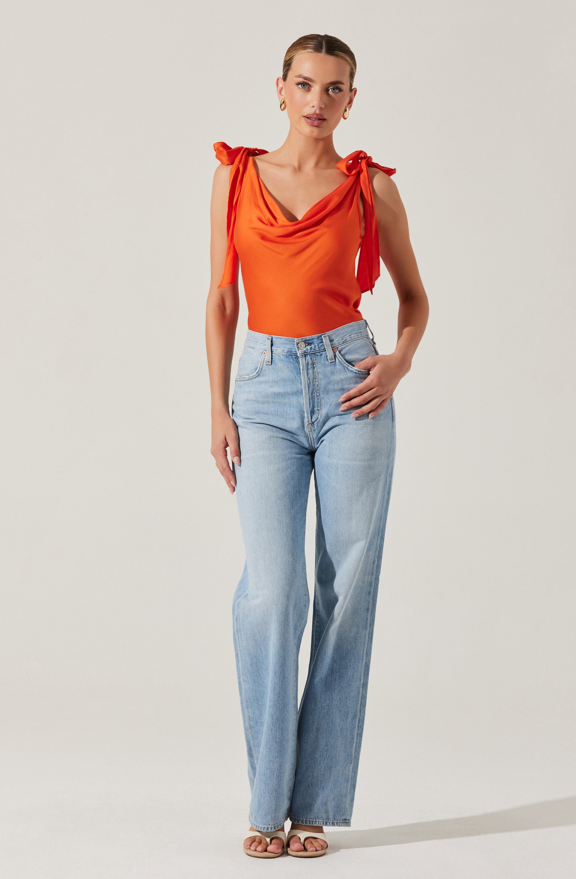 A woman is wearing an orange sleeveless top with a draped neckline and shoulder straps tied into bows on each shoulder. She pairs the top with high-waisted, light-wash, wide-leg jeans.