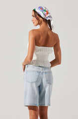 A woman is wearing a white, strapless, ruched top with an elasticized, smocked design at the waist. She is also wearing light-wash denim shorts with a relaxed fit and rolled-up hems. Additionally, she has a colorful, floral-patterned headscarf tied around her head.