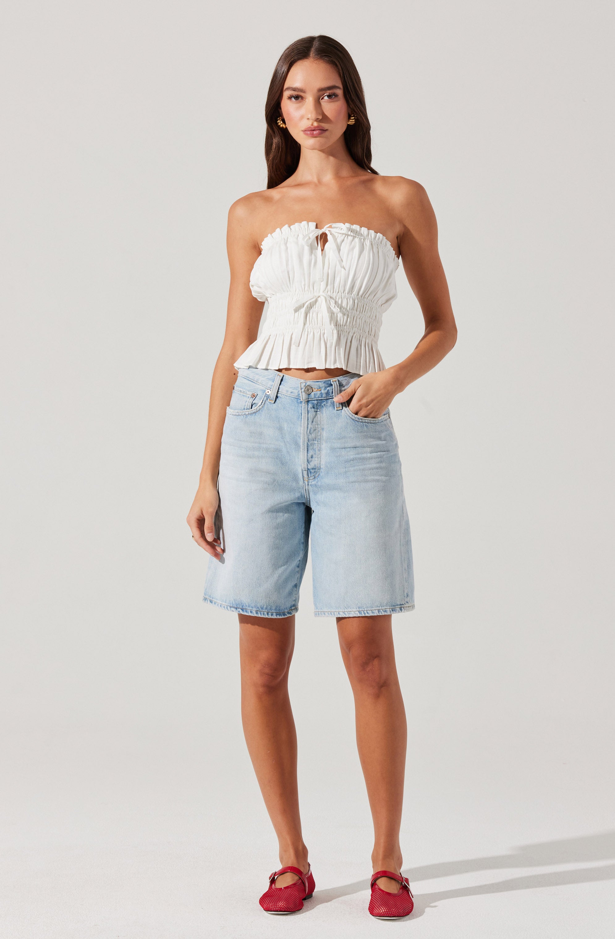 A woman is wearing a strapless, white, ruffled crop top with a gathered, elasticized waist and a small keyhole opening at the front. She is also dressed in light blue, knee-length denim shorts with a classic five-pocket design. She is wearing red flat shoes with a bow detail.