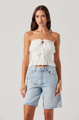 A woman is wearing a strapless, white, gathered crop top with ruffled hem and a small tie at the center. She is also dressed in light-wash denim shorts that reach above the knees, with a classic five-pocket design and a silver button closure.