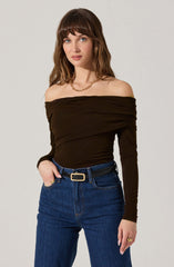 A woman is wearing an off-the-shoulder, long-sleeved black top that is ruched and fitted. She pairs it with high-waisted dark blue jeans and a black belt with a gold rectangular buckle. She accessorizes with a gold chain necklace and a ring on her left hand.