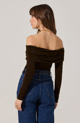 A woman is wearing an off-the-shoulder black top with gathered fabric around the neckline and sleeves. She has paired the top with high-waisted blue jeans. She is accessorized with a gold chain necklace and a ring on her left hand.