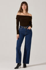 A woman is wearing a dark brown, off-the-shoulder top with ruched detailing. She has paired it with high-waisted, straight-leg blue jeans with a black belt that has a rectangular gold buckle. She completes the look with black heeled boots.