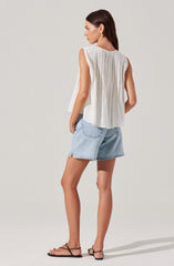 A woman is wearing a sleeveless, white, pleated top with a loose fit and a round neckline. She pairs it with light blue denim shorts that have a relaxed fit and rolled hems. She completes the outfit with black flat sandals that have thin straps.