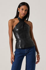 A woman is wearing a sleeveless black leather top with a halter neck design that features a twisted fabric detail at the neck, leaving her shoulders and arms bare. She pairs the top with high-waisted blue jeans. She accessorizes with silver earrings, a silver bracelet, and rings.