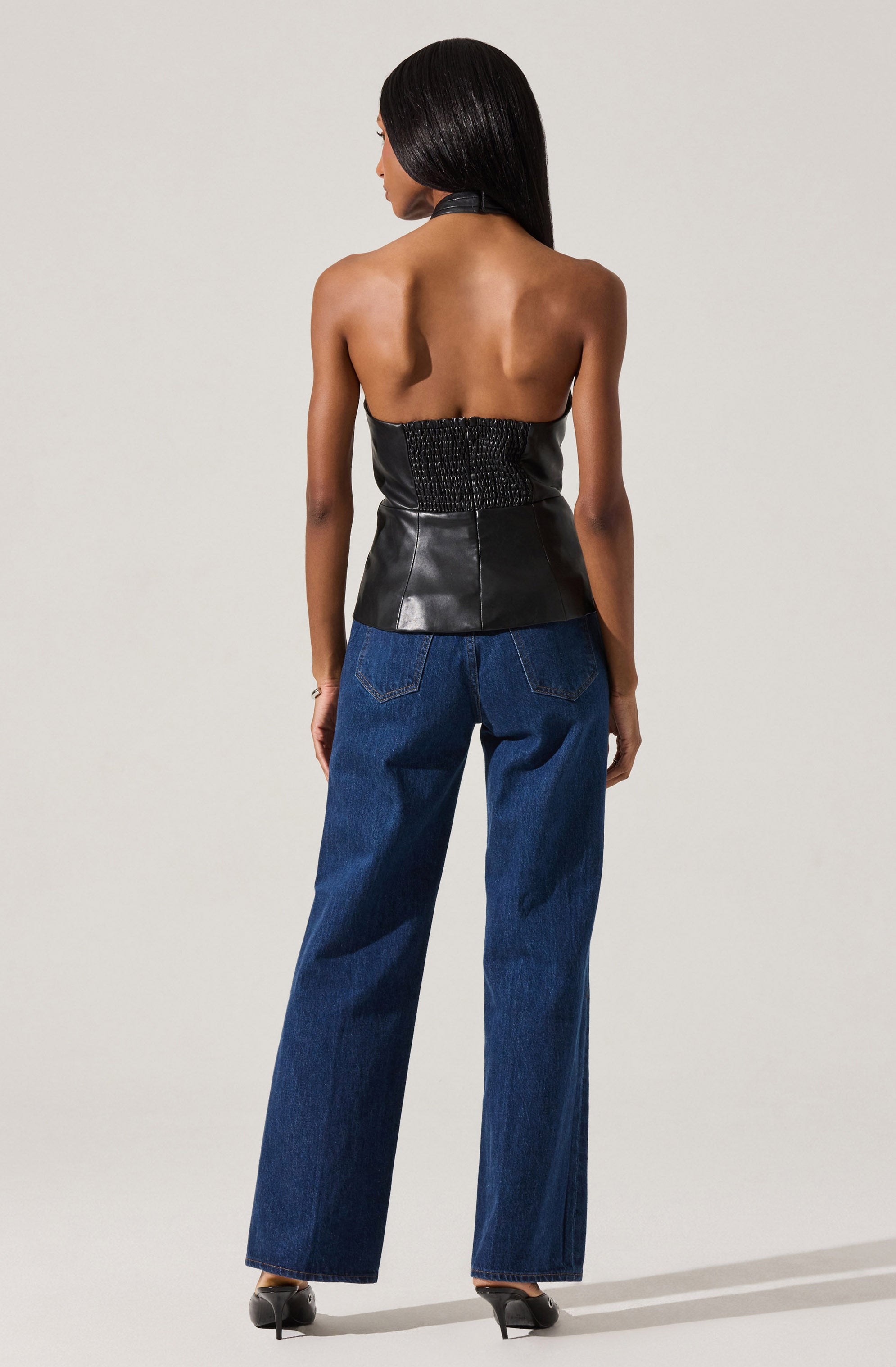 A woman is wearing a sleeveless, strapless black leather top with textured details on the back. She pairs it with high-waisted, wide-leg blue jeans and black high-heeled shoes.