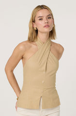 A woman is wearing a beige sleeveless halter top with a draped neckline that crosses at the collarbone. The top has a button-down front with four visible buttons and a small slit at the bottom hem. She pairs it with light-colored pants.