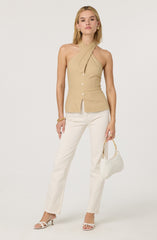 A woman is wearing a beige sleeveless top with a twisted halter neckline, featuring a front button closure. She pairs it with high-waisted, straight-leg white trousers. She accessorizes with gold jewelry, including earrings and rings, and is holding a white woven handbag with a gold handle. She completes her look with white heeled sandals.