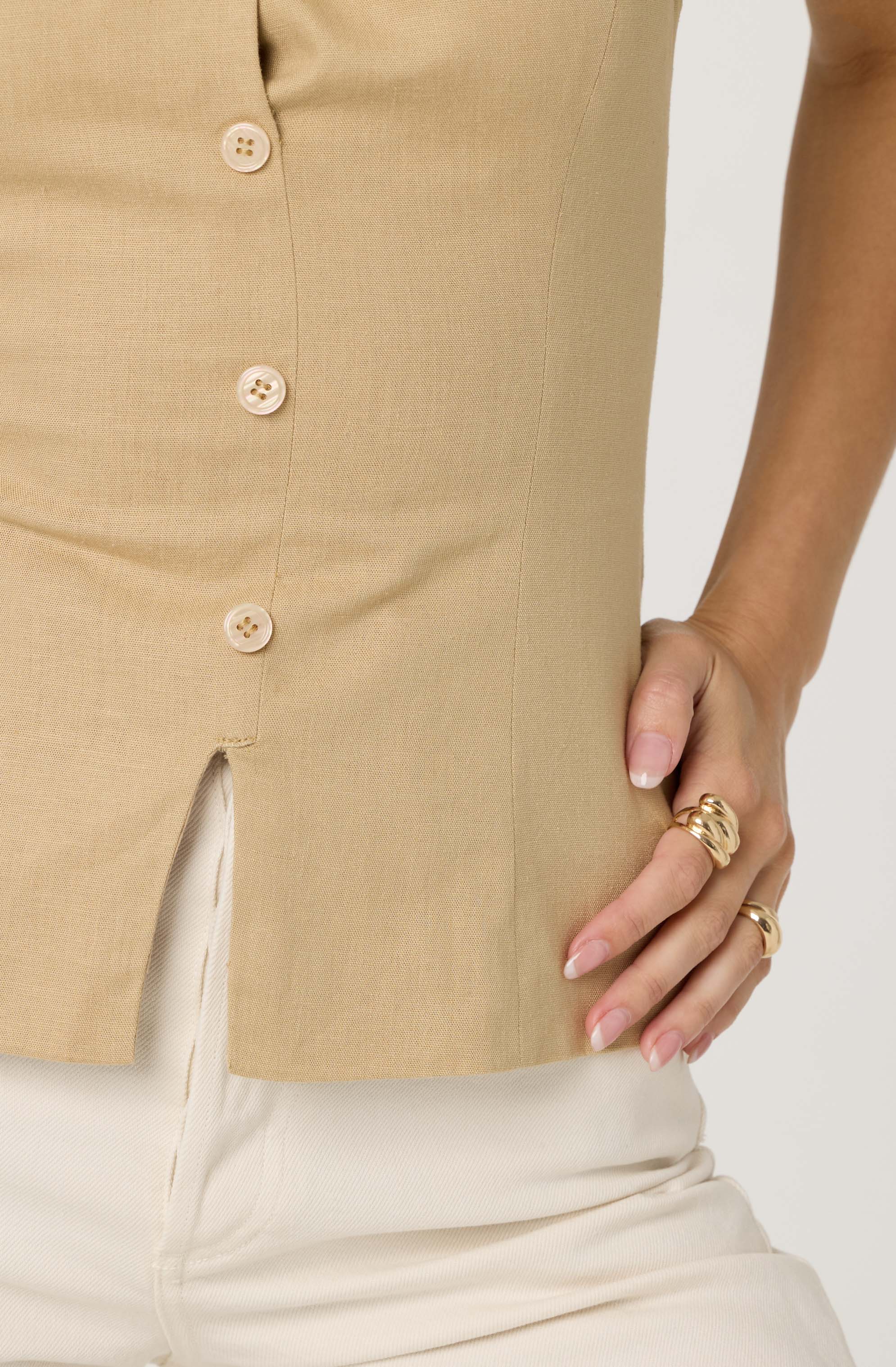 A person is wearing a beige sleeveless button-up top with four light-colored buttons. The top has a small slit at the hem in the front. They are also wearing light-colored pants and have their right hand resting on their hip, adorned with multiple gold rings.