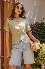 A woman is wearing a short-sleeved, light green, button-up blouse with a round neckline and decorative gold-tone buttons down the front. She pairs it with high-waisted, light-wash denim shorts and accessorizes with round, dark sunglasses, gold earrings, and a crocheted straw handbag.