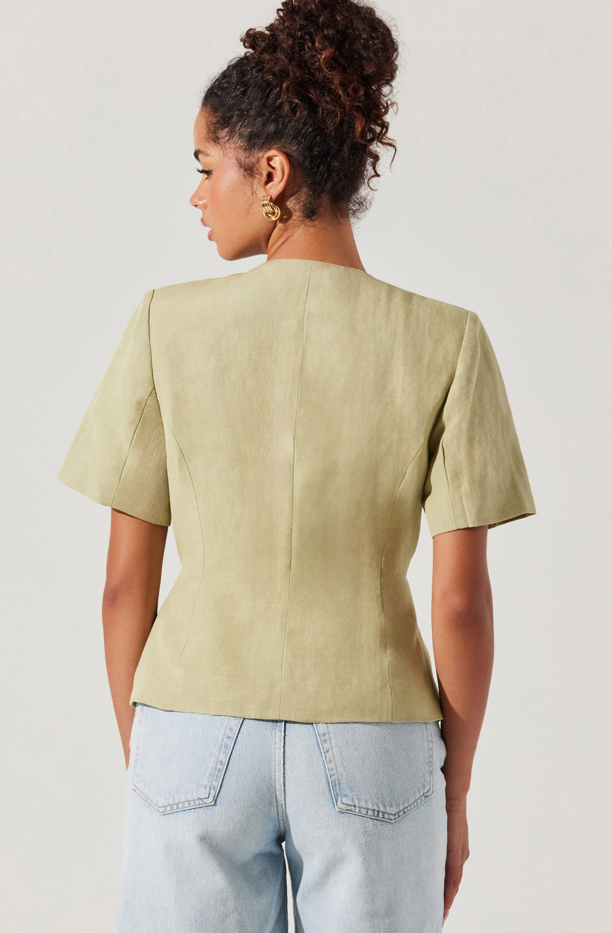 The image shows a woman wearing a light green, short-sleeved blazer with a structured fit and seam details, paired with light blue denim jeans. The blazer has a smooth, slightly textured appearance. She has dark curly hair styled in a high puff and is accessorized with gold hoop earrings.