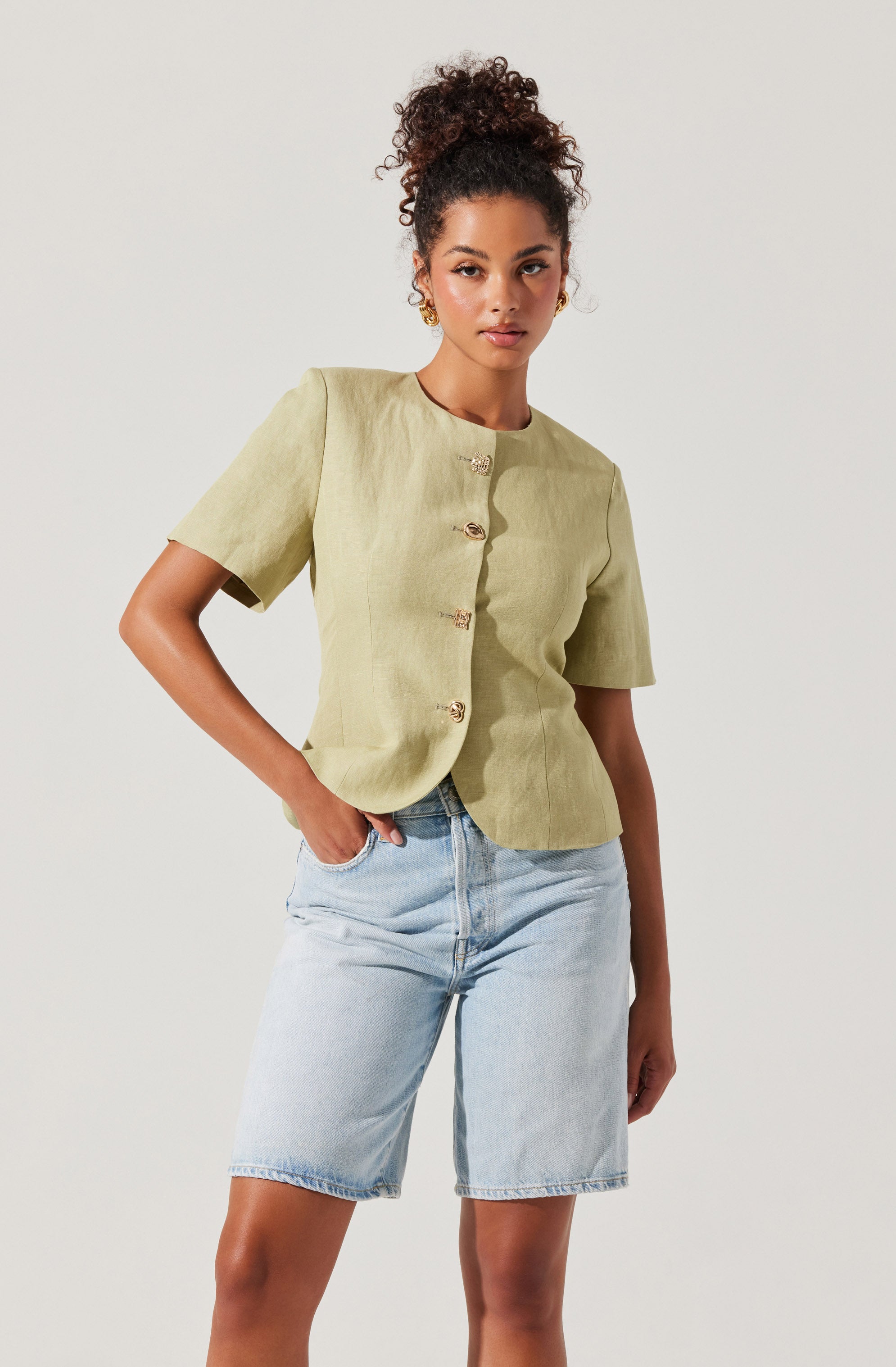 A woman is wearing a light green, short-sleeved blouse with a round neckline and decorative buttons down the front. She pairs it with light blue denim shorts that have a relaxed fit and a high waist.