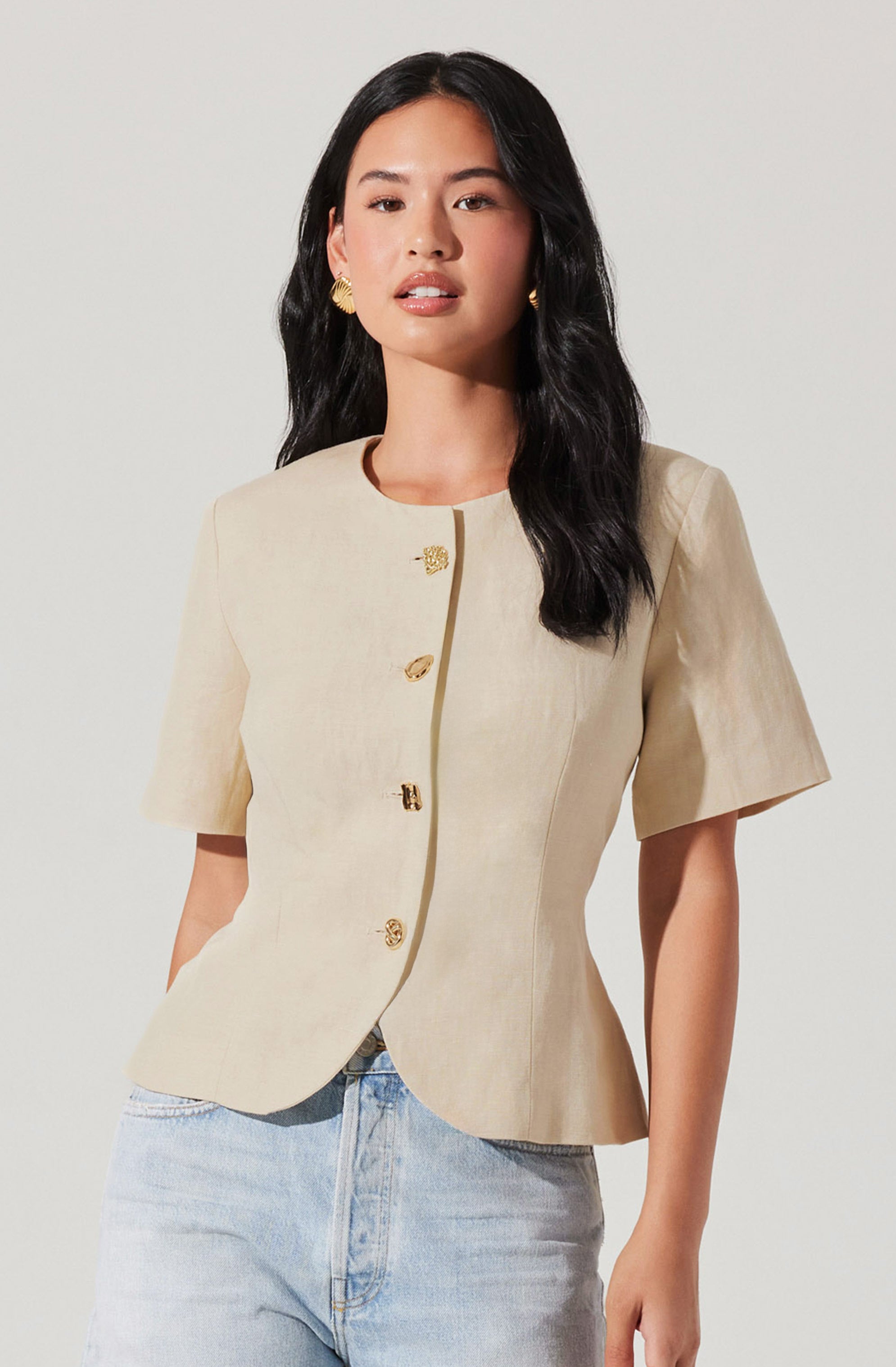 A woman is wearing a beige, short-sleeved blazer with a rounded neckline and decorative gold buttons down the front. She has paired it with light-wash jeans.