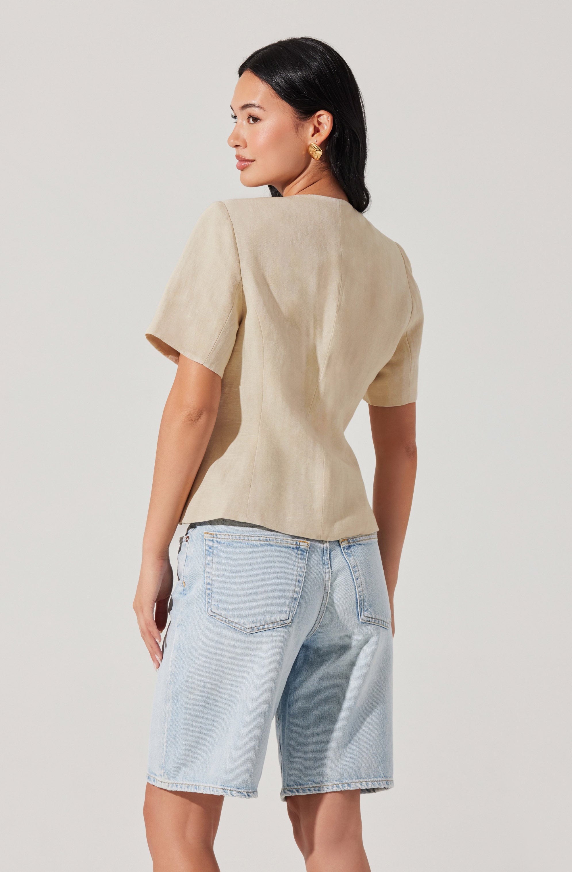 A woman is wearing a beige short-sleeve top with a simple, elegant design and light-wash denim shorts with a relaxed fit. The top has a round neckline and a slightly textured fabric, while the shorts feature visible stitching and back pockets. She has black hair and is accessorized with gold earrings.