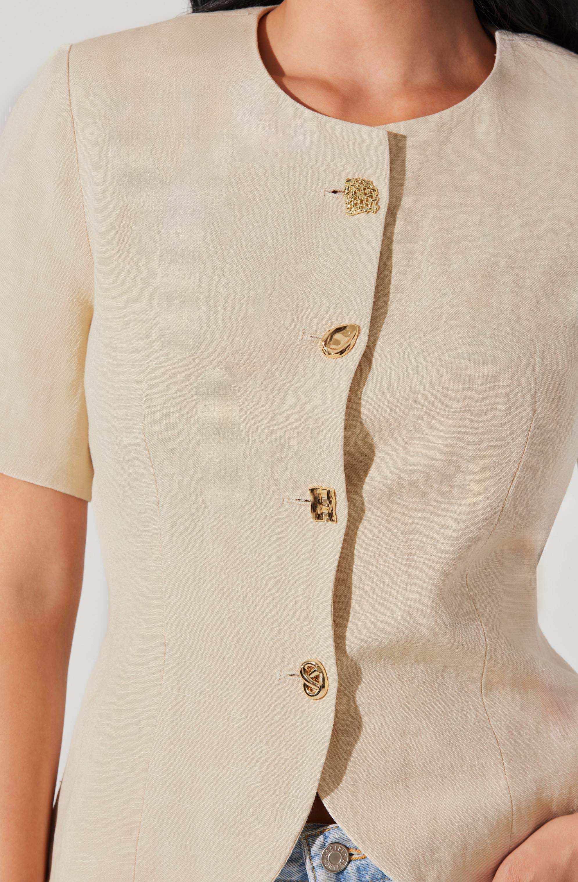 A close-up of a woman's beige short-sleeve jacket with a round neckline, featuring five decorative gold-tone buttons down the front. The buttons have various designs, including textured and smooth finishes.