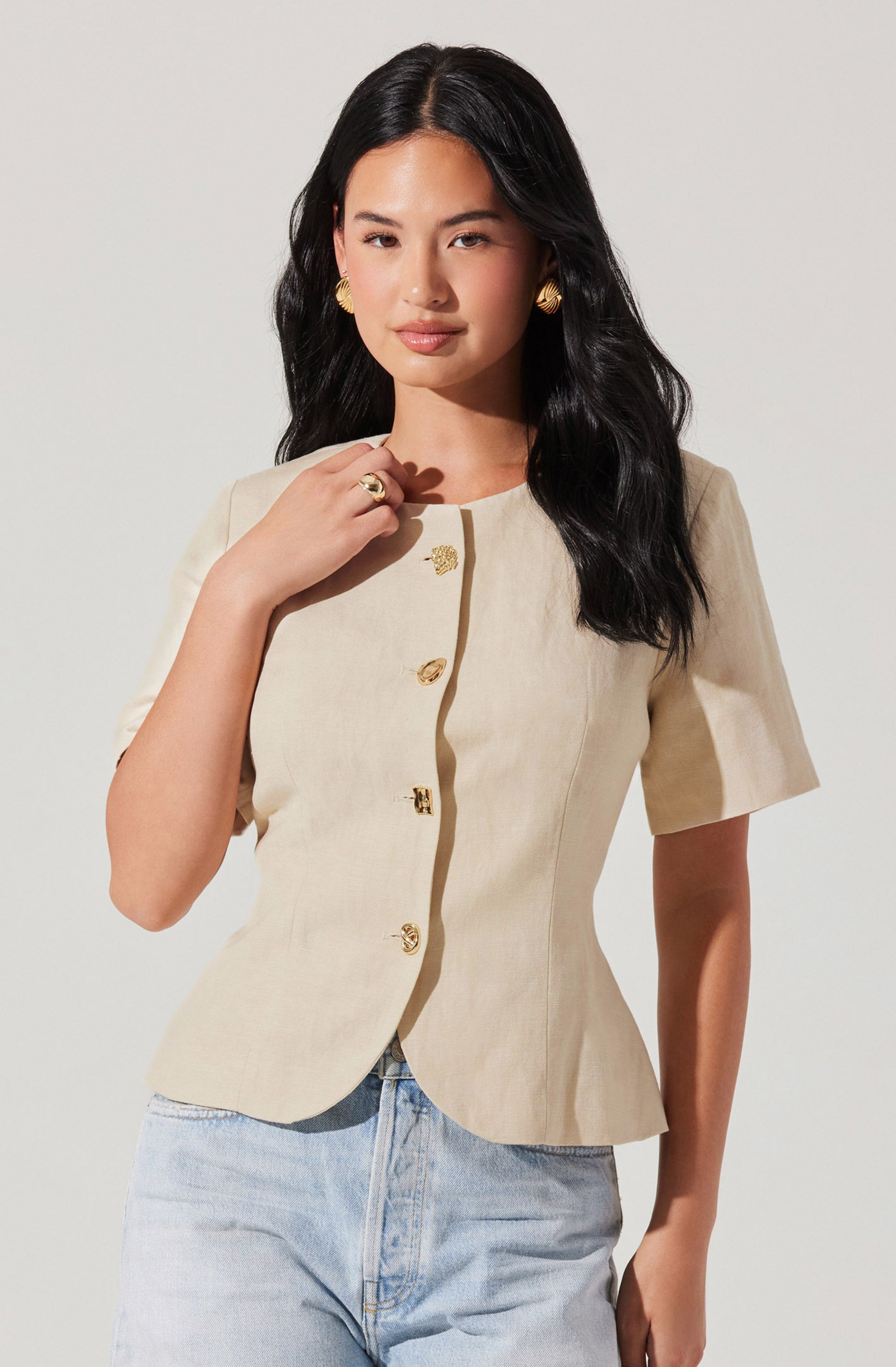 A woman is wearing a beige short-sleeve blazer with a round neckline and decorative gold buttons down the front. She has paired it with light-wash jeans and accessorized with large gold earrings and a gold ring.