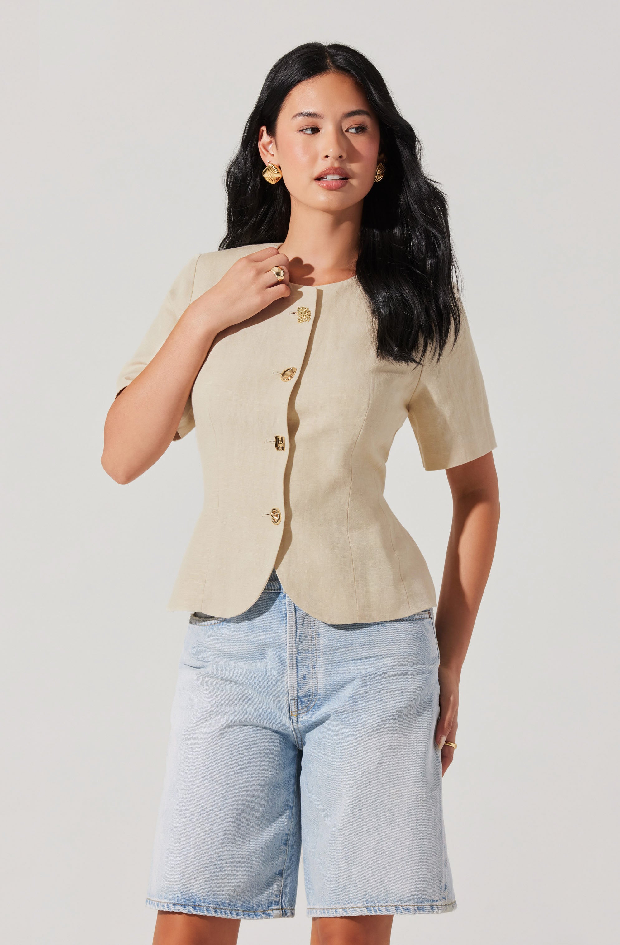 A woman is wearing a beige short-sleeved jacket with gold buttons down the front and gold earrings. She also has a gold ring on her right hand and is dressed in light-wash denim shorts.