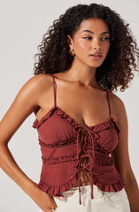 A woman is wearing a rust-colored, sleeveless crop top with thin spaghetti straps. The top features ruffled trim along the neckline, hem, and horizontal seams, with a front tie closure made of matching fabric. She pairs it with light-colored pants.