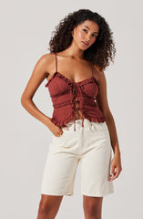 A woman is wearing a dark red, sleeveless top with thin spaghetti straps and ruffle details along the neckline, hem, and around the waist. The top also has a ruched front with adjustable ties in the center. She pairs it with high-waisted, off-white knee-length shorts.