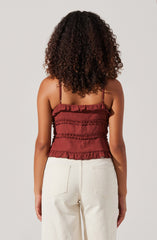 A woman is wearing a sleeveless, rust-colored top with thin spaghetti straps. The top features multiple horizontal ruffled layers that add texture and visual interest. She pairs it with high-waisted, off-white pants.