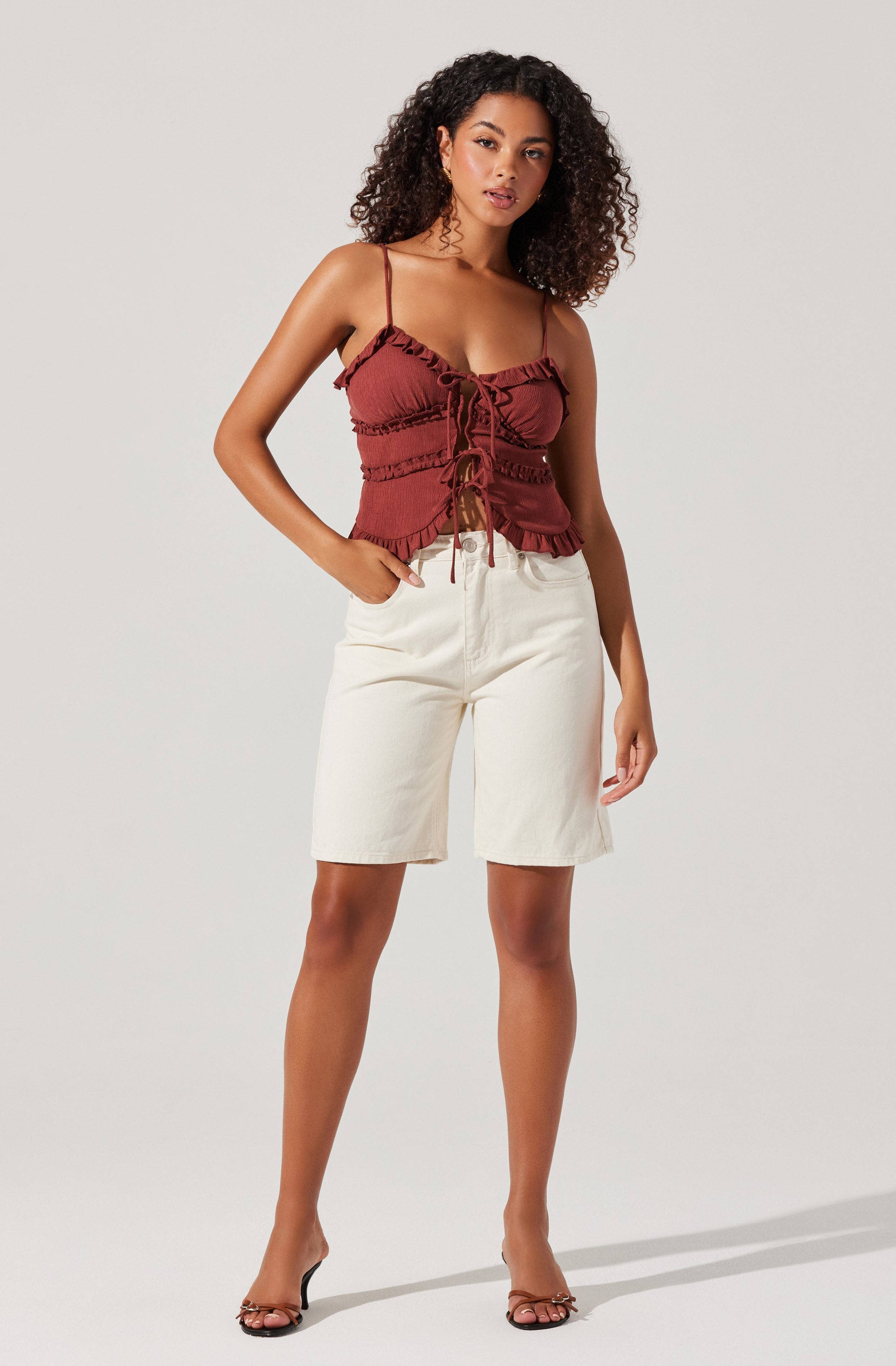 A woman is wearing a reddish-brown, ruffled, sleeveless top with thin spaghetti straps and a front tie detail. She pairs it with high-waisted, off-white knee-length shorts and black open-toe heels.