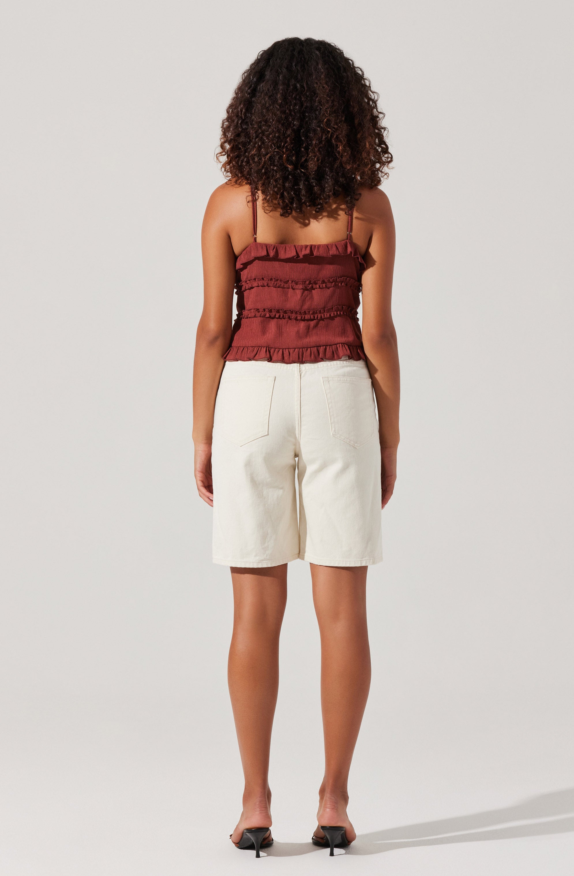 A woman is wearing a sleeveless, ruffled, reddish-brown top with thin spaghetti straps and horizontal ruffle details. She is also wearing high-waisted, loose-fitting cream-colored shorts with visible back pockets. The outfit is completed with black open-toe heeled sandals.