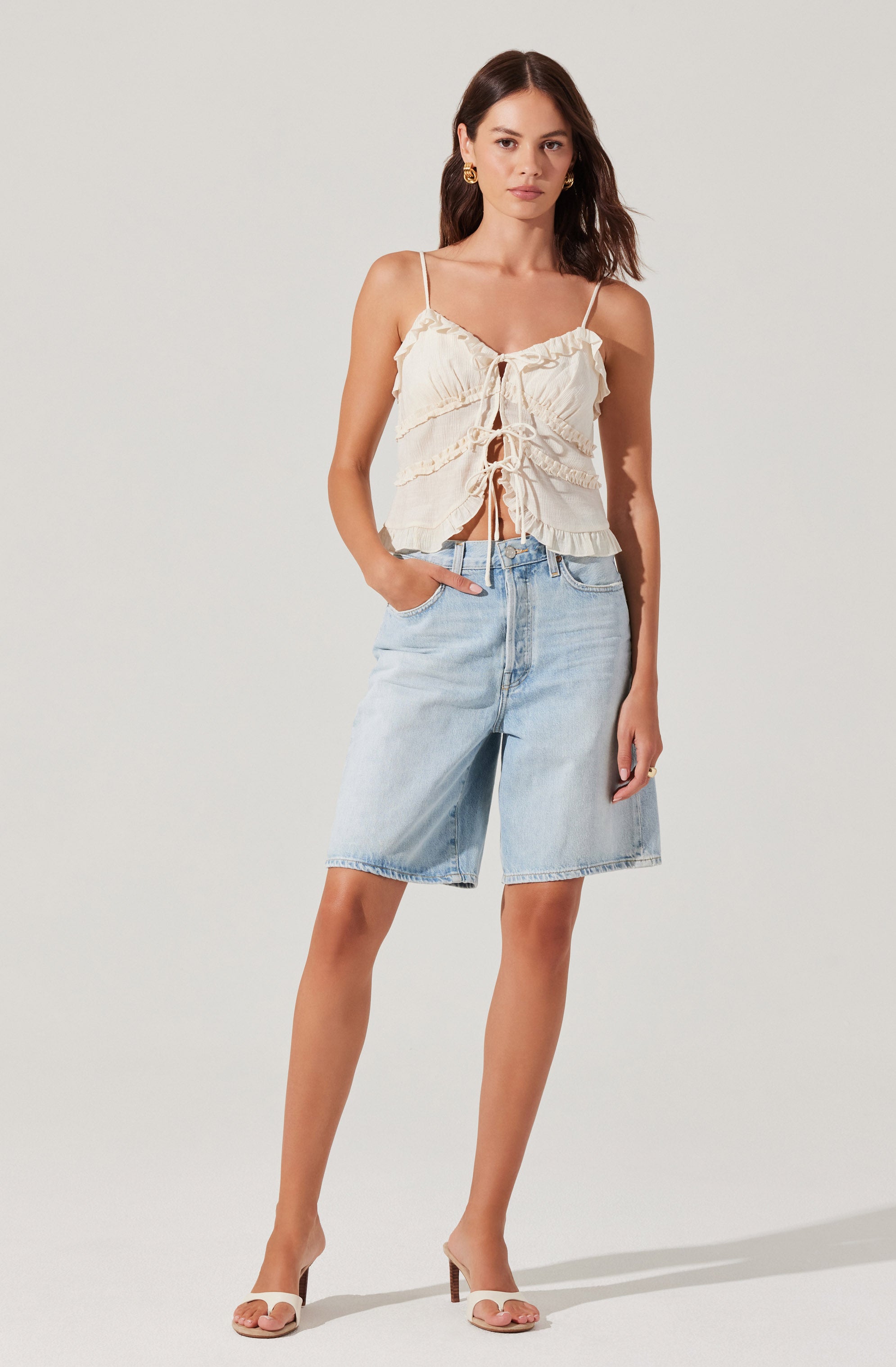 A woman is wearing a cream-colored, sleeveless, ruffled top with spaghetti straps and a front tie detail. She has paired the top with light blue, knee-length denim shorts. She is also wearing beige open-toe high-heeled sandals and gold hoop earrings.