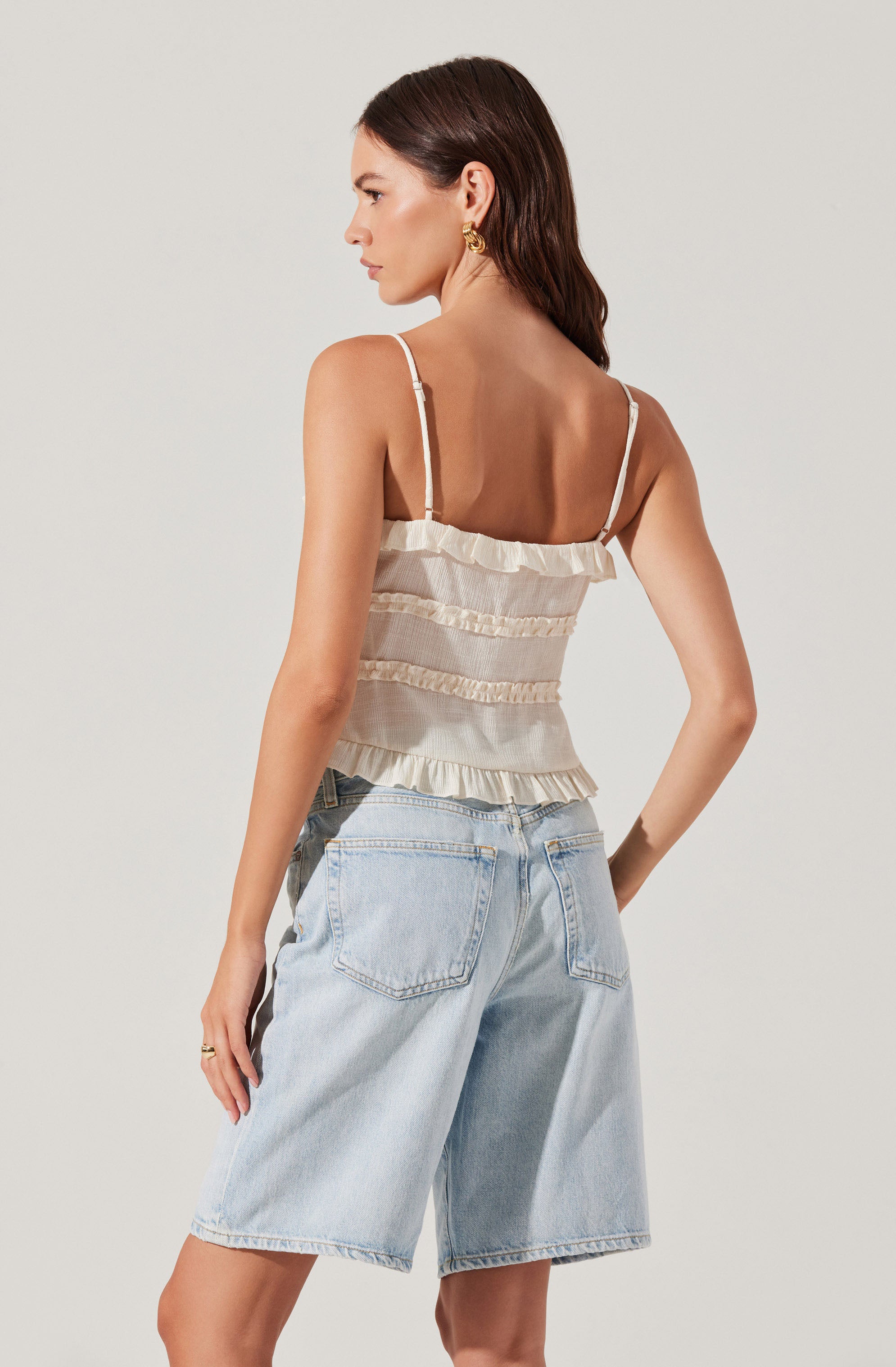 A woman is wearing a sleeveless, cream-colored, ruffled crop top with thin spaghetti straps and light-wash, high-waisted denim shorts with large back pockets.