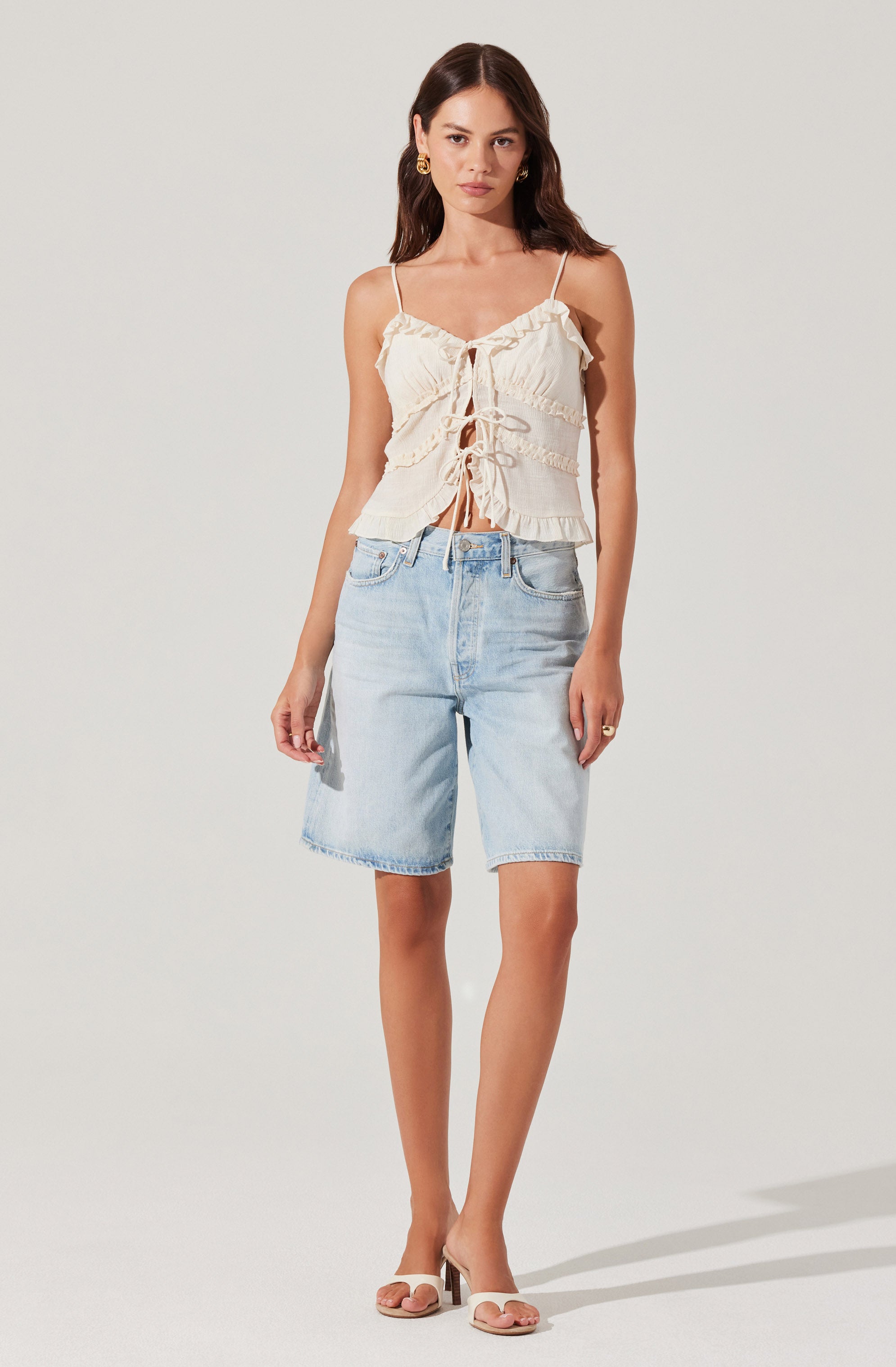 A woman is wearing a cream-colored, ruffled, sleeveless top with spaghetti straps and a front tie detail. She is paired with light wash denim Bermuda shorts that are knee-length. She completes the outfit with beige open-toed heels and gold earrings.