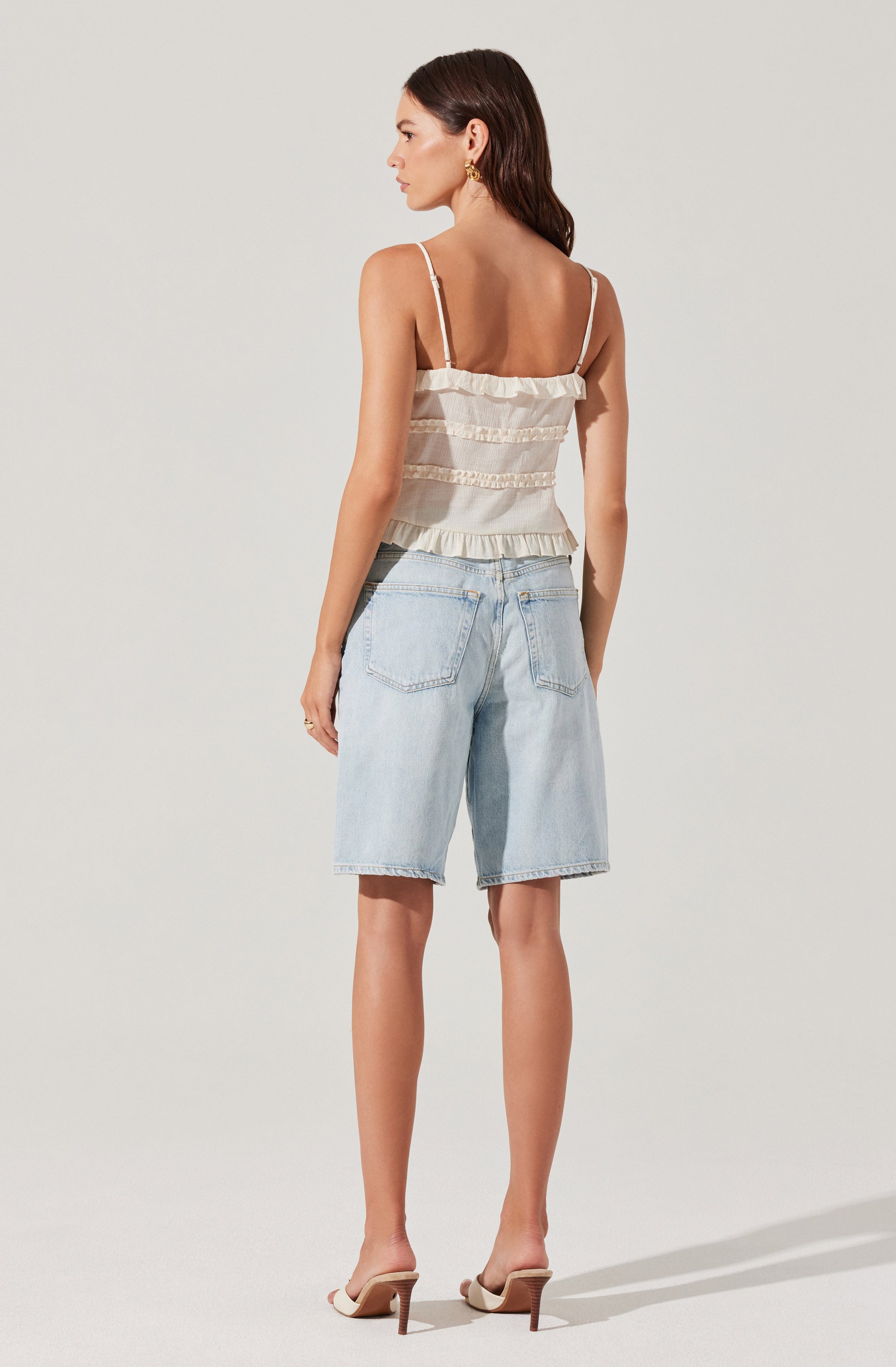 A woman is wearing a cream-colored sleeveless top with ruffled horizontal stripes and thin shoulder straps. She pairs it with light blue high-waisted denim shorts that have a relaxed fit and rolled cuffs. She is also wearing nude high-heeled sandals.