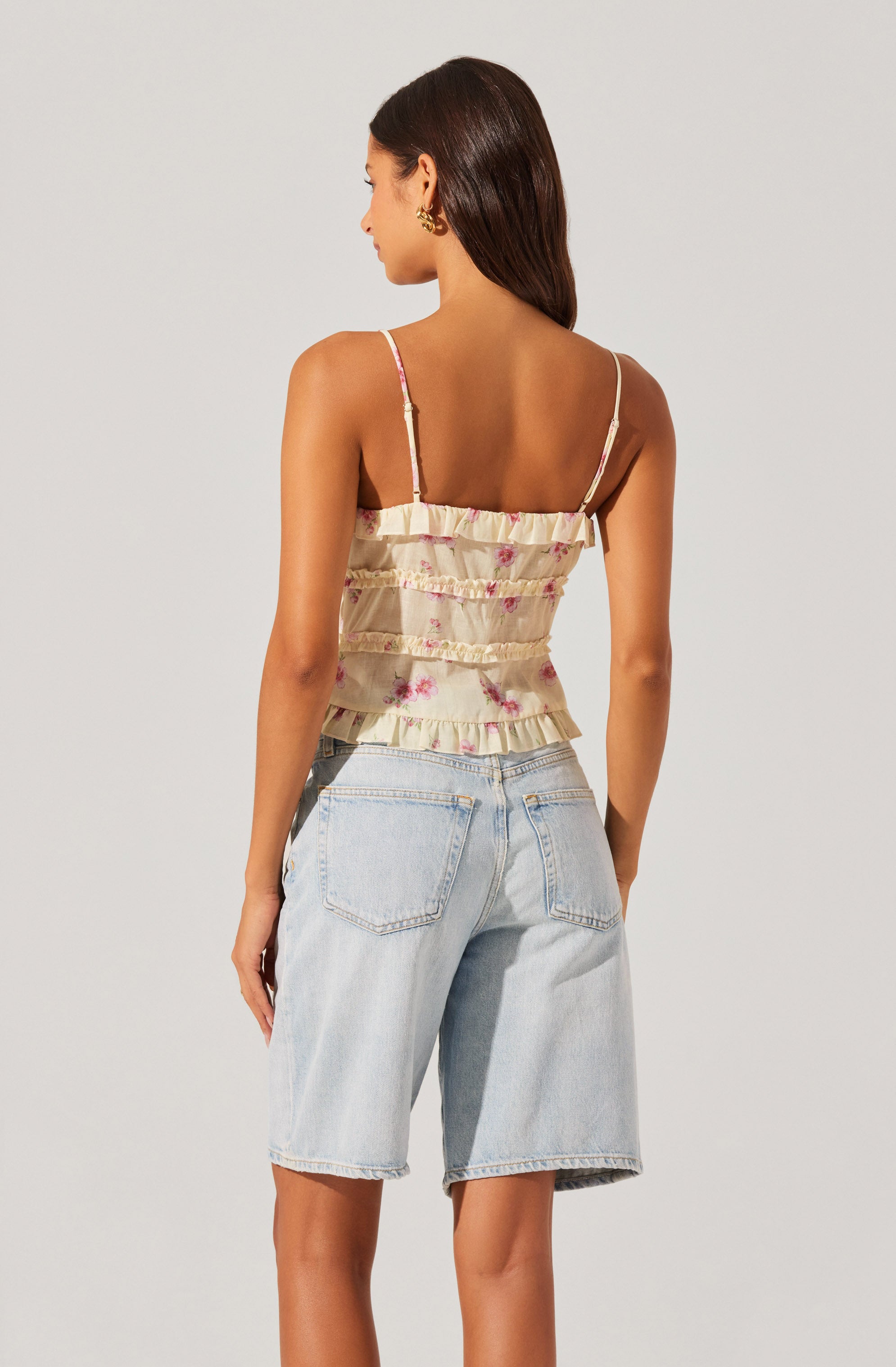 A woman is wearing a sleeveless, ruffled top with thin spaghetti straps. The top has a light, cream-colored background with pink floral patterns. She is also wearing light-wash, high-waisted denim shorts with large back pockets.