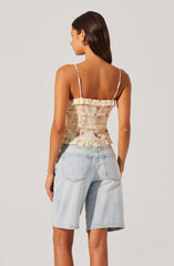 A woman is wearing a sleeveless, ruffled top with thin spaghetti straps. The top has a light, cream-colored background with pink floral patterns. She is also wearing light-wash, high-waisted denim shorts with large back pockets.