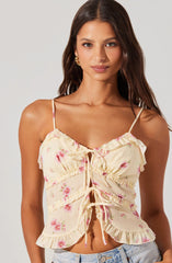 A young woman is wearing a light yellow, floral-patterned, sleeveless top with thin spaghetti straps. The top features ruffles along the edges and a keyhole neckline tied with a bow at the center. The fabric has pink and green floral prints, and the top has a slightly ruffled hem.