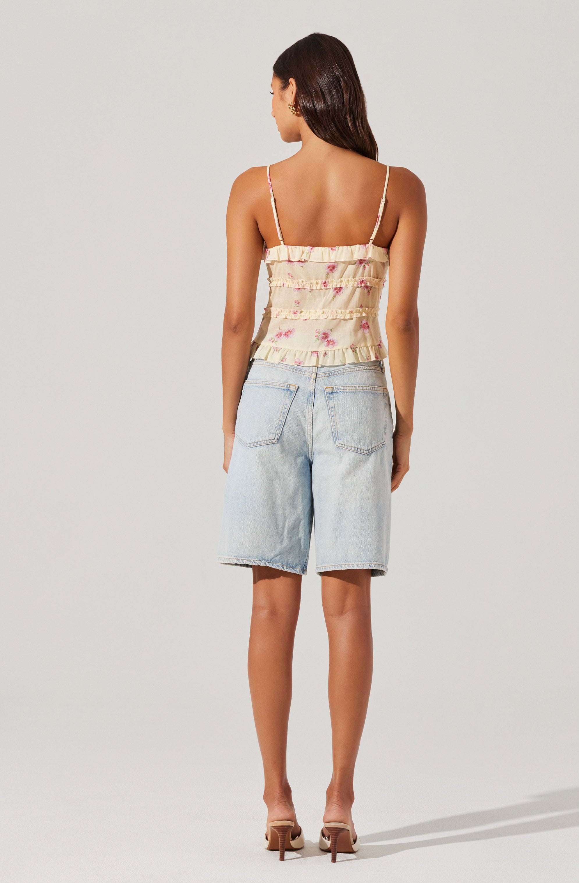 A woman is wearing a sleeveless, ruffled, floral-printed top with thin spaghetti straps. She is also wearing light-wash, knee-length denim shorts with large back pockets and a frayed hem. She has on nude-colored high-heeled shoes.