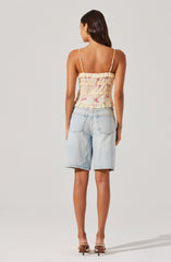 A woman is wearing a sleeveless, ruffled, floral-printed top with thin spaghetti straps. She is also wearing light-wash, knee-length denim shorts with large back pockets and a frayed hem. She has on nude-colored high-heeled shoes.