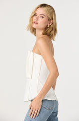 A young woman with shoulder-length blonde hair is wearing a strapless white top with pleated detailing in the center and a slightly flared hem. She is also wearing light blue denim jeans. The background is plain and light-colored.
