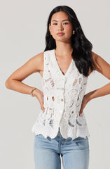A woman is wearing a sleeveless white lace top with intricate cutout patterns and scalloped edges. The top features a button-down front and a scalloped hemline. She pairs it with light blue jeans.