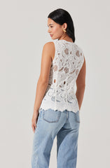 A woman is wearing a sleeveless white lace top with a floral and swirl pattern, featuring a scalloped hem. She is also wearing light blue, high-waisted jeans with visible stitching on the back pockets.