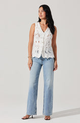 A woman is wearing a sleeveless, white, lace top with intricate cut-out patterns and scalloped edges. She pairs it with light-wash, wide-leg jeans and black high-heeled sandals.