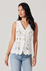 A woman is wearing a sleeveless white crochet top with a scalloped hem and intricate openwork patterns, paired with light blue denim jeans.