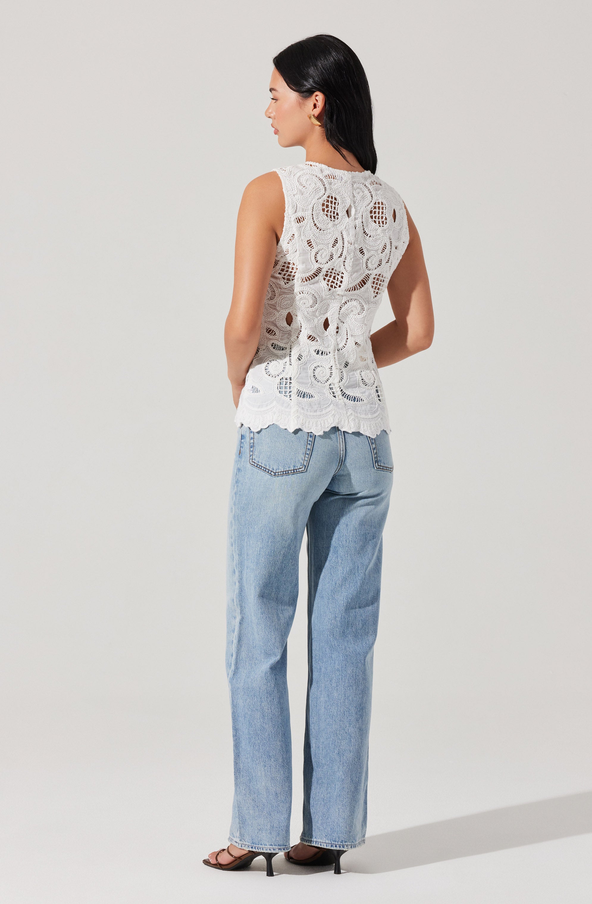 A woman is wearing a sleeveless white lace top with intricate circular and floral patterns, featuring scalloped edges at the bottom. She pairs it with light blue high-waisted jeans and black high-heeled sandals.
