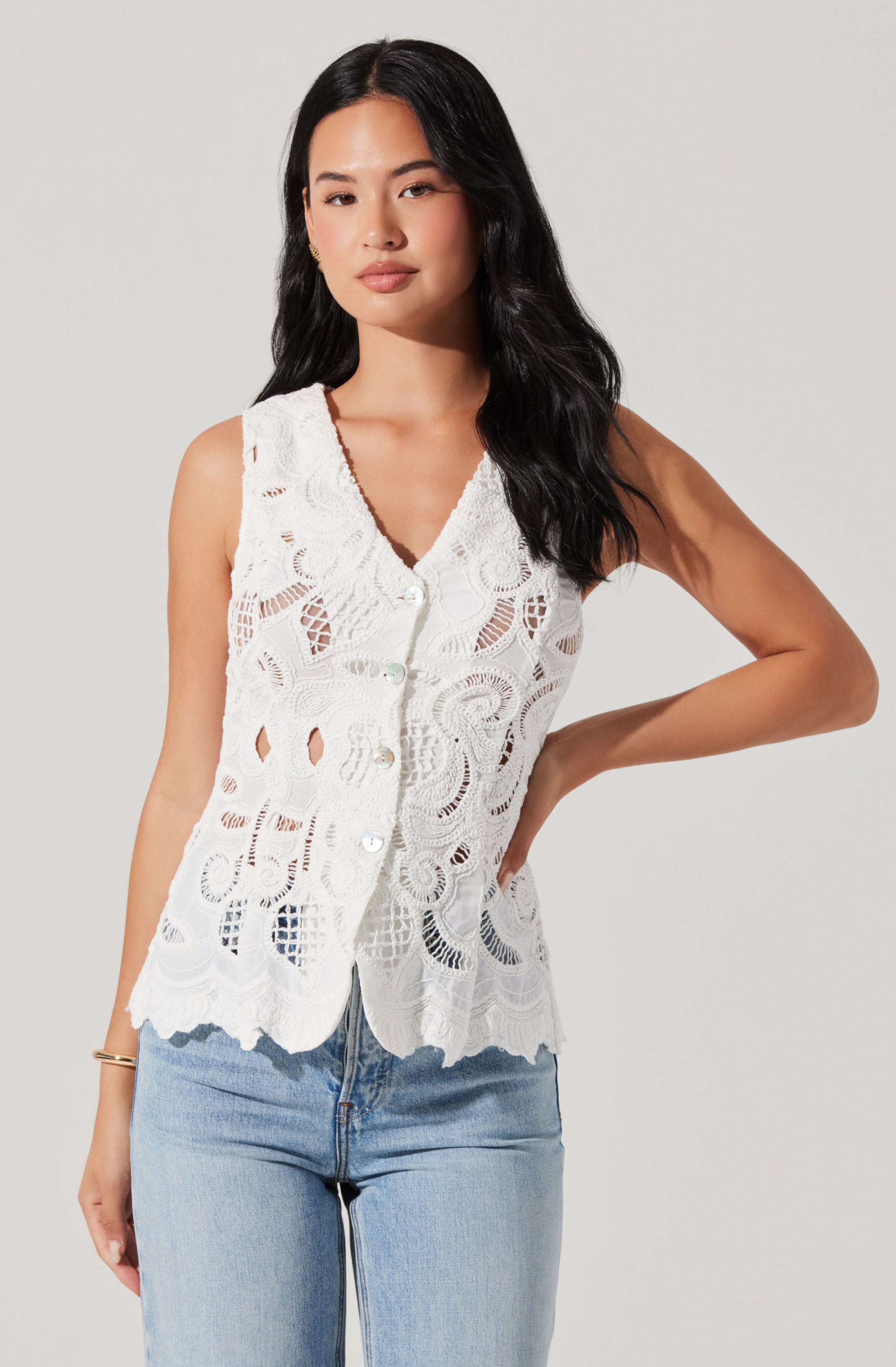 A woman is wearing a sleeveless, white openwork lace top with a scalloped hem and a button-down front. She pairs it with light blue jeans.