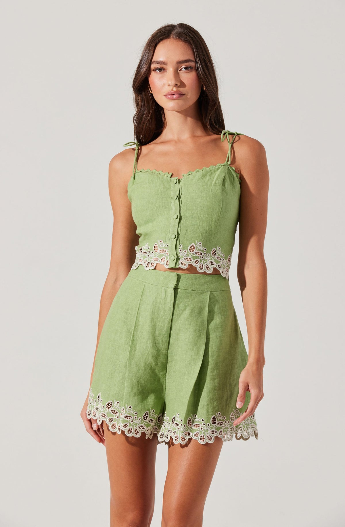 A woman is wearing a matching light green sleeveless top and shorts set. The top has thin tie straps and a scalloped neckline, with a button-down front. Both the top and shorts feature decorative white eyelet embroidery along the hemlines.