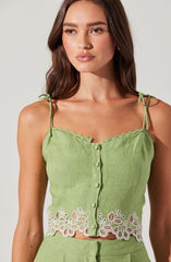 A woman is wearing a sleeveless, light green top with thin, tie-up shoulder straps. The top features a scalloped neckline and a row of buttons down the front. The lower part of the top has intricate white floral embroidery with cut-out details.