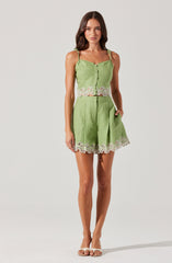 A woman is wearing a matching light green two-piece outfit consisting of a sleeveless, button-front crop top with ruffled straps and lace trim along the hem, and high-waisted shorts with pleats and lace trim at the hem. She is also wearing open-toe sandals.