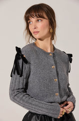 A woman is wearing a gray knitted cardigan with gold heart-shaped buttons down the front. The cardigan features black ribbon bows on the shoulders. She is also wearing a black leather skirt and gold jewelry, including hoop earrings and rings.